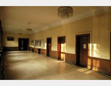 Langer Flur in einem historischen Gebäude mit hellbeigen Wänden, hohen Decken und mehreren Holztüren auf der rechten Seite. Der Raum ist mit großen Fenstern auf der linken Seite sowie gläsernen Kronleuchtern an der Decke ausgestattet und wird von warmem Tageslicht durchflutet.