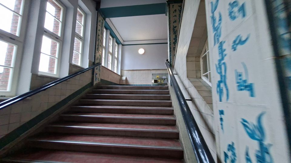Innenansicht eines Treppenhauses mit breiter Treppe und schwarzen Handläufen, flankiert von hohen Sprossenfenstern auf der linken Seite. Die Wände sind teils gekachelt und zeigen blaue Schriftzeichen bzw. Verzierungen, oben führt die Treppe zu einem hellen Flur mit runder Deckenleuchte.