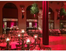 Ein festlich dekorierter Veranstaltungsraum mit runden, gedeckten Tischen, Kerzenleuchtern und umgedrehten, hängenden Weihnachtsbäumen an der Decke. Im Hintergrund sind rote Vorhänge und große Fensterbögen sichtbar, die auf eine elegante Atmosphäre hinweisen.
