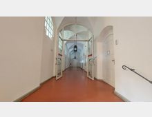 Langer, heller Korridor mit gewölbter Decke und zwei geöffneten Glastüren in der Mitte. Der Boden ist rot, an den Wänden befinden sich weiße Türen sowie schmale Fenster, und schwarze Laternen hängen von der Decke.