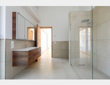 Modernes Badezimmer mit großformatigen, beigen Fliesen, einer gläsernen Dusche, einem hängenden Holzwaschtisch mit integriertem Spiegel und Zugang zu einem angrenzenden, hellen Flur durch eine geöffnete Holztür.