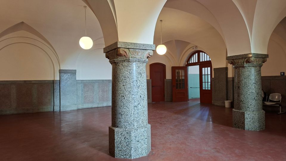 Gewölbte Eingangshalle mit großen Granitsäulen und Rundbogen-Decke, hängenden Kugelleuchten sowie rotbraunem Boden und grauer Wandverkleidung. Im Hintergrund befinden sich Holztüren mit Glaseinsätzen und ein hoher Rundbogen-Durchgang.