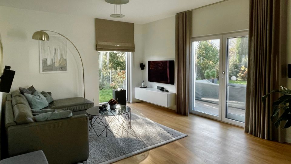 Helles, modernes Wohnzimmer mit grauem Ecksofa, Teppich und rundem Couchtisch, dazu eine Bogen-Stehlampe und ein wandmontierter Fernseher über einem weißen Lowboard. Große Glas-Terrassentüren mit Vorhängen und ein weiteres Fenster führen in einen Gartenbereich; Holzboden und neutrale Wandgestaltung prägen den Raum.