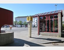 Straßenszene mit Taxi-Wartesäule und gelbem Taxi-Schild in der Mitte, umgeben von einem modernen Wartehäuschen mit roten Rahmen und einer stark befahrenen Straße im Hintergrund. Im Hintergrund sind ein historisches weißes Gebäude und mehrere parkende Autos sichtbar.