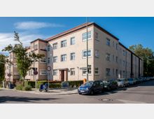 Wohngebäude aus der Nachkriegszeit mit drei Stockwerken und beiger Fassade an einer Straßenecke, umgeben von geparkten Autos und vereinzelten Bäumen. Ein Straßenschild mit der Aufschrift "Monopolstraße" sowie Verkehrsschilder sind sichtbar.