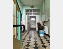 Flur in einem älteren Gebäude mit schwarz-weiß kariertem Boden, grünen Wänden und hohen Decken. Am Ende des Flurs befindet sich eine geöffnete Doppeltür mit Glaselementen, die zu einem weiteren Bereich mit einer Holz-Eingangstür und einem halbkreisförmigen Fenster darüber führt.