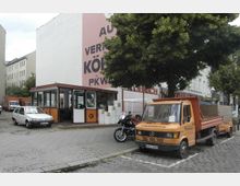 Ein kleiner Betriebshof mit einer orange-schwarzen Bürohütte, auf der ein Schriftzug mit Autovermietungsangeboten prangt. Davor stehen ein weißer Kleinwagen, ein orangefarbener Transporter mit Anhänger und ein Motorrad, umgeben von Bäumen und Wohnhäusern im Hintergrund.