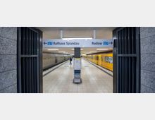 U-Bahn-Station mit gelbem Zug auf der rechten Seite und einem Mittelbahnsteig. Ein Schild weist Fahrtrichtungen der Linie U7 zu "Rathaus Spandau" links und "Rudow" rechts aus.