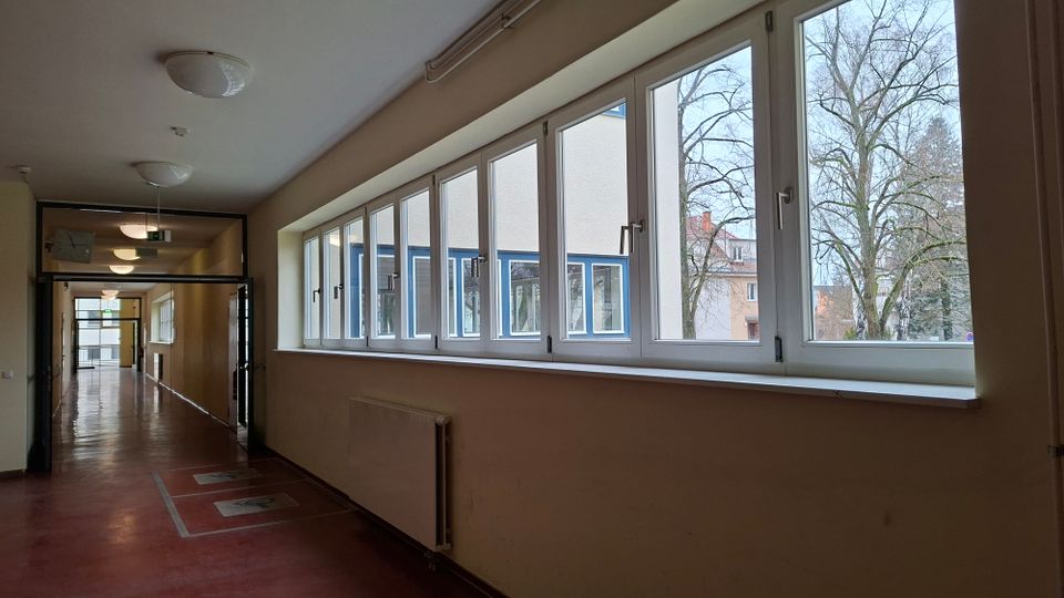 Langer Innenflur eines öffentlichen Gebäudes mit rotem Boden, Deckenleuchten und einer Reihe großer Fenster entlang der rechten Seite. Durch die Fenster sind ein gegenüberliegendes Gebäude sowie Bäume im Außenbereich zu sehen.