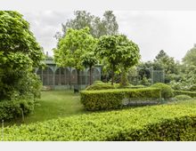 Ein gepflegter Garten mit geschnittenen Hecken, kleinen Bäumen und einer Rasenfläche. Im Hintergrund steht ein gläserner Pavillon mit grünen Metallelementen und Ziegelwand sowie ein dekorativer Torbogen.