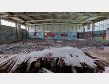 Innenraum einer verlassenen, heruntergekommenen Halle mit beschädigtem Holzboden, offenen Lücken und zerfallenem Holz. Die Wände sind mit Graffiti bedeckt, und das Tageslicht fällt durch große, fensterartige Glasblöcke an den Seitenwänden.