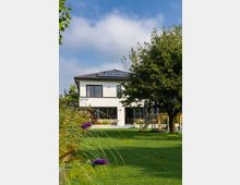 Modernes zweistöckiges Wohnhaus mit weißer Fassade und großen Fenstern, umgeben von einem gepflegten Garten mit Rasen, Bäumen und Blumen; auf dem Dach sind Solarmodule sichtbar. Im Vordergrund stehen ein Baum und lila Blüten, daneben ein Sitzbereich mit Gartenmöbeln nahe der Terrasse.