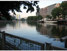 Flusslandschaft mit Promenade: Ein ruhiger Fluss mit angelegten Booten am rechten Ufer, gesäumt von modernen Gebäuden und Bäumen. Im Vordergrund ist ein Geländer mit Blick auf das Wasser zu sehen, und im Hintergrund stehen mehrstöckige Wohn- und Geschäftsgebäude.