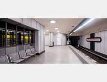 U-Bahn-Station mit schlichtem, modernem Design, weißen Wandverkleidungen und Deckenleuchten. Auf der rechten Seite das Stationsschild „Bismarckstraße“, links Metallbänke und ein Fenster, dahinter ein vorbeifahrender gelber U-Bahn-Zug.