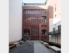 Ein Innenhof mit einem rot verklinkerten Gebäude im Hintergrund, das schmale, hohe Fenster und Rundbögen aufweist. Der Hof ist mit Kies und Pflastersteinen gestaltet, umgeben von Sitzbänken und kleinen Grünflächen, mit modernen Gebäudeelementen auf der rechten Seite.