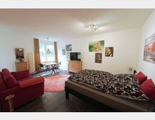 Wohnraum mit Doppelbett, rotem Sofa, Esstisch und Stühlen vor einem großen Fenster mit beigen Vorhängen. An den Wänden hängen mehrere Gemälde, und ein Fernseher steht auf einer Kommode; ein runder, roter Teppich liegt in der Mitte des Raumes.