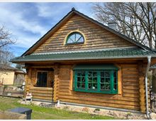 Ein Holzhaus aus massiven Baumstämmen mit grünem Dach und einem großen, grünen Erkerfenster. Im Vordergrund sind ein kleiner Garten mit Steinen und Pflanzen sowie ein offener Eingangsbereich zu sehen.