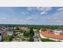 Stadtansicht mit roten Ziegeldächern und einer Mischung aus Wohnhäusern und begrünten Flächen, im Hintergrund ein Wasserturm und ausgedehnte Waldlandschaft unter blauem Himmel mit wenigen Wolken.
