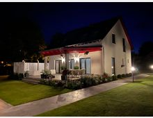 Einfamilienhaus mit weißer Fassade und rotem Dach, umgeben von einem gepflegten Garten mit Rasen und Blumenbeeten, bei Nacht beleuchtet durch Gartenlampen; eine Terrasse mit Sitzmöbeln und Blumentöpfen liegt im Vordergrund.