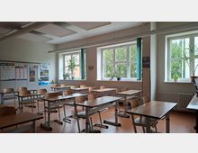 Heller Klassenraum mit mehreren Reihen aus Holztischen und Stühlen sowie großen Fenstern an der Längsseite mit grünen Vorhängen und Pflanzen auf den Fensterbänken. An den Wänden hängen Unterrichtsplakate, und an der Decke sind rechteckige Akustikpaneele und Leuchtleisten montiert.