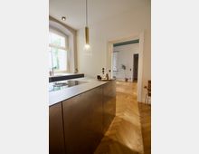 Moderne Küchenzeile mit einer glatten metallischen Front und einer Hängelampe mit Glas- und Goldelementen, daneben ein großes Fenster mit weißem Rahmen, das Tageslicht hereinlässt. Der Raum hat einen warmen Holzfußboden im Fischgrätenmuster, der in einen angrenzenden, hellen Raum mit minimaler Einrichtung führt.