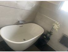 Modernes Badezimmer mit freistehender weißer Badewanne vor grauen Wandfliesen und dunklem Boden, ergänzt durch eine kleine Ablage mit Pflanze. Ein Fenster sorgt für Tageslicht, daneben sind ein Handtuchhalter und Armaturen an der Wand zu sehen.