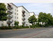 Mehrstöckiges Wohngebäude in heller Putzfassade mit Balkonen, umgeben von Bäumen und Grünflächen. Im Vordergrund eine Straße mit Gehweg und einigen parkenden Autos.