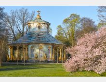 Pavillon im Rokoko-Stil mit runder Struktur, grünlicher Fassade und goldenen Verzierungen, umgeben von Bäumen und einem blühenden rosa Kirschbaum im Vordergrund.