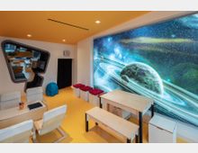 Moderner Kinder- oder Lernraum mit gelbem Boden und orangefarbener Decke, möbliert mit Holztischen, weißen Stühlen und Hockern mit roten Sitzkissen. Eine große Wandmalerei zeigt eine Darstellung des Weltalls mit Planeten und Sternen.