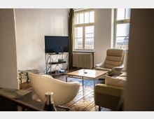 Wohnzimmer mit Holzboden und schlichtem Interieur, bestehend aus mehreren Sesseln, einem Holztisch und einem Fernseher auf einem Metallregal. Große Fenster lassen Tageslicht herein, ein Heizkörper befindet sich unter den Fenstern.