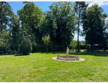 Grüne Parklandschaft mit Wiese und Hintergrund aus hohen Bäumen. Im Zentrum steht ein dekorativer Brunnen mit einer Statue, die eine menschliche Figur darstellt, umgeben von weißem Kies.