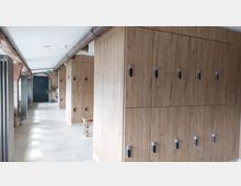 Ein Flur mit hellen Bodenfliesen, Holzspinden an der rechten Seite und einer Backsteinwand links. Im Hintergrund ist eine dunkle Tür mit einem Schild sowie Pflanzen in Töpfen sichtbar.