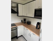 Moderne Küche mit weißen Schränken und Arbeitsplatte aus Holz. Zu sehen sind ein eingebauter Backofen, ein Kochfeld, eine Mikrowelle, eine Kaffeemaschine, ein Wasserkocher und ein Toaster.