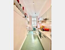 Schmale Küche mit pastellfarbenen Wänden und grünem Bodenbelag. Auf der rechten Seite befinden sich moderne Küchenschränke, Herd und Spüle, auf der linken Seite Regale mit Gewürzen und eine Waschmaschine; ein Fenster mit Tageslicht und ein kleiner Essbereich mit einem Stuhl befinden sich am Ende des Raumes.