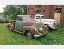 Zwei alte, rostige Pickup-Trucks stehen auf einer grünen Wiese vor einem roten Backsteingebäude mit bogenförmigen Fenstern. Im Hintergrund ist ein dritter Wagen vor einem Baum sichtbar.