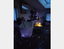 Ein modernes Badezimmer mit freistehender Badewanne, umgeben von schwachem, punktuellem Licht, das einen sternenähnlichen Effekt erzeugt. Pflanzen, ein Dachfenster und ein kleines Holztablett mit Kerzenlicht schließen die gemütliche Szene ab.