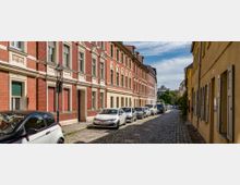 Eine schmale, gepflasterte Straße mit geparkten Autos auf beiden Seiten, flankiert von einer gelben und einer roten Backsteinfassade historischer Wohnhäuser mit verzierten Fensterrahmen. Im Hintergrund sind Bäume und vereinzelte moderne Gebäude zu erkennen, unter einem bewölkten Himmel.