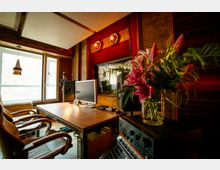 Ein modernes Tonstudio mit einem großen Holztisch, Ledersesseln und technischem Equipment wie Bildschirmen und Mischpulten. Links befindet sich ein Fenster mit natürlichem Lichteinfall, während ein Glasvasenstrauß mit Lilien und Farnen auf einem Gerät in der rechten vorderen Ecke steht.