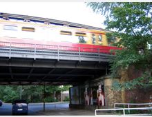 Eine rot-gelbe S-Bahn fährt über eine erhöhte Stahlbrücke, die von Backsteinpfeilern gestützt wird. Unter der Brücke befinden sich eine Straße, parkende Autos und Werbeplakate, umgeben von Bäumen und Stadtgrün.