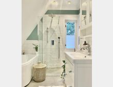 Modernes Badezimmer mit weiß gefliesten Wänden, einer begehbaren Dusche mit Glaswand und Regenduschkopf, einer freistehenden Badewanne sowie einem weißen Waschtisch mit Spiegel. Ein Korb und grüne Pflanzen dienen als Dekoration.