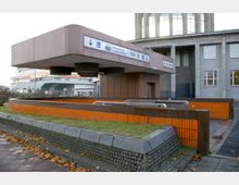 Eingang zu einer unterirdischen Haltestelle oder Passage mit braun-orangefarbenen Wänden und Geländern. Im Hintergrund sind ein modernes Gebäude, ein Plakat und ein Wegweiser mit Hinweisen auf das Internationale Congress Centrum (ICC), den ZOB und weitere Verbindungen zu sehen.