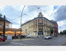 Straßenszene mit einer historischen Ecke aus drei Gebäuden: ein gelb-rotes, verziertes Wohnhaus mit Balkonen, daneben ein orangefarbenes Gebäude mit einem Café und gelben Sonnenschirmen im Außenbereich. Straßenbahnschienen und Stromleitungen verlaufen durch die Szene, umgeben von Autos und Fußgängern.