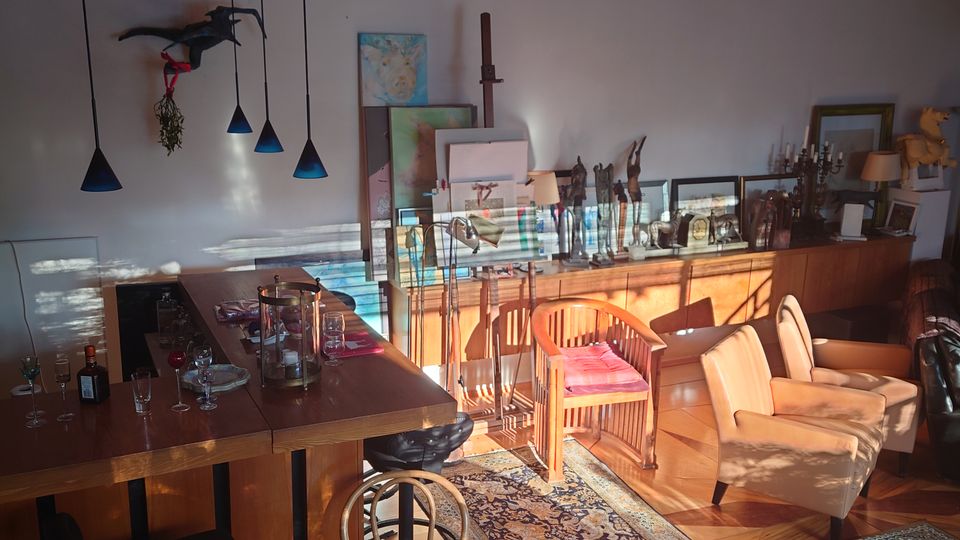 Wohnraum mit Holzbalkendecke und dekorativem Oberlicht aus buntem Glas. Der Raum enthält eine Bar mit Hockern, gemütliche Sitzmöbel, Teppiche und verschiedene Kunstgegenstände sowie Dekorationen.