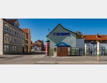 Straßenszene mit einer Bushaltestelle, öffentlichen Toiletten und einem Gebäude mit Werbeschild für Autoreparaturen. Im Hintergrund stehen Wohnhäuser mit Ziegeldächern und Fassaden im älteren Stil.