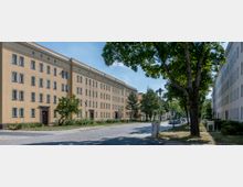 Mehrstöckige Wohngebäude mit cremefarbenen Fassaden und regelmäßiger Fensteranordnung säumen eine ruhige, mit Kopfsteinpflaster belegte Straße. Auf beiden Straßenseiten stehen Bäume, die Schatten spenden, und im Hintergrund sind parkende Autos und Laternen erkennbar.