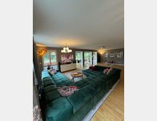 Großes, modernes Wohnzimmer mit einer L-förmigen, dunkelgrünen Samtcouch, die mit bunten Kissen dekoriert ist. Der Raum hat helle Holzböden, große Fenster mit Blick ins Grüne, eine minimalistische TV-Wand und eine auffällige Deckenleuchte in Kugelform.