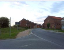 Straßenszene mit einer Kurve, flankiert von historischen Backsteingebäuden mit roten Ziegeldächern; ein Gehweg verläuft parallel zur Straße, und links befindet sich eine grasbewachsene Fläche mit Verkehrszeichen. Der Himmel ist leicht bewölkt.