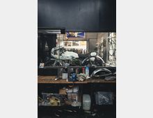 Ein Werkstattbereich mit einer hölzernen Werkbank im Vordergrund, auf der Werkzeuge, Ölbehälter und Stofflappen liegen. Im Hintergrund hinter einer Glasscheibe steht ein weißes Motorrad, umgeben von Fahrrädern und Wanddekorationen.