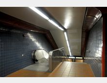 Unterirdischer U-Bahn-Eingang mit blauen Wandfliesen, einer Rolltreppe und Fliesenboden. Die Decke ist mit langen, schlanken Leuchtstofflampen beleuchtet.