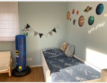Ein Kinderzimmer mit hellblauen Wänden, dekoriert mit Wandstickern von Planeten und kleinen Sternen. Links steht ein aufblasbarer blauer Boxsack mit Batman-Motiv, daneben ein Bett mit blauer Bettwäsche und einem Kuscheltier, sowie eine weiße Lamellenjalousie am Fenster.